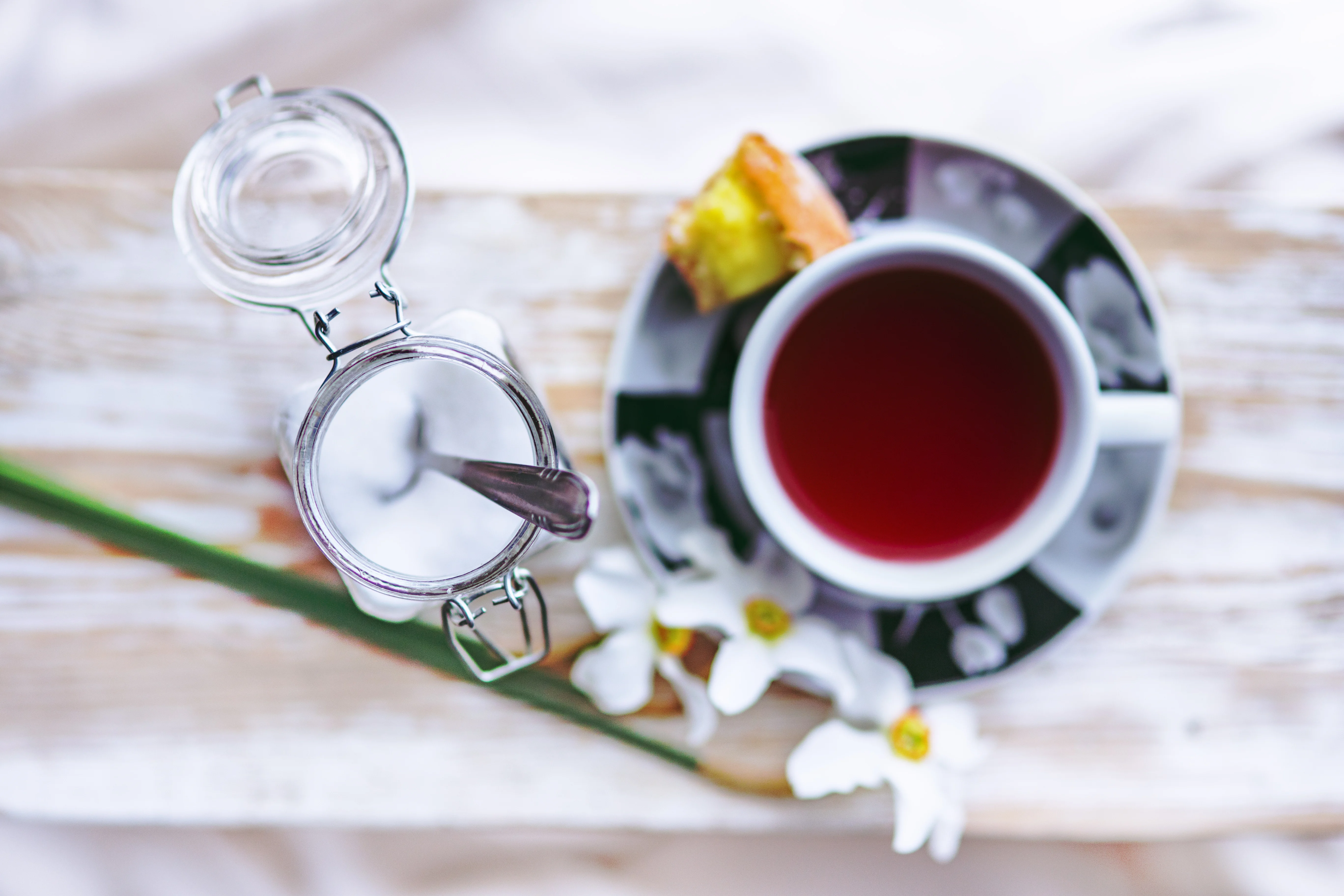 cup of tea with flowers and a pastrey on the rim of the dish and a glass of sugar next to it